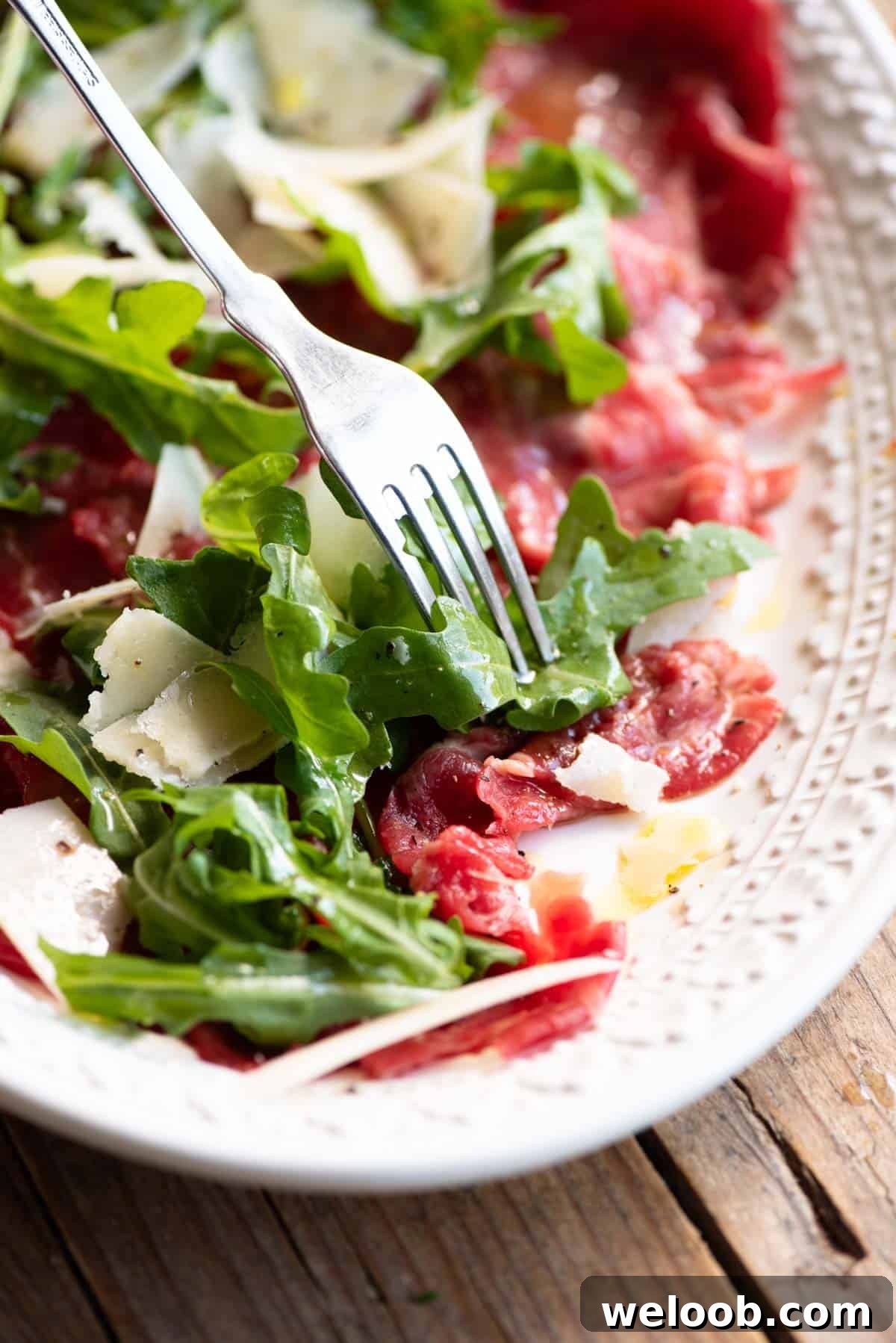A close up of beef carpaccio, arugula and Grana Padano cheese on a plate with a fork.