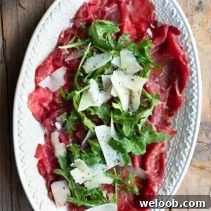 A close up cropped image of beef carpaccio on a white serving plate with arugula and shavings of Grana Padano cheese.