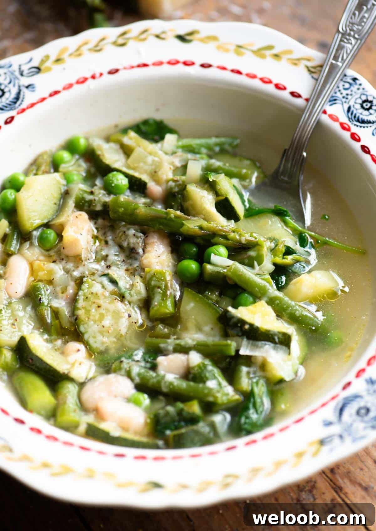 A close up of a green vegetable soup in a patterned bowl with asparagus, peas, zucchini and green beans.