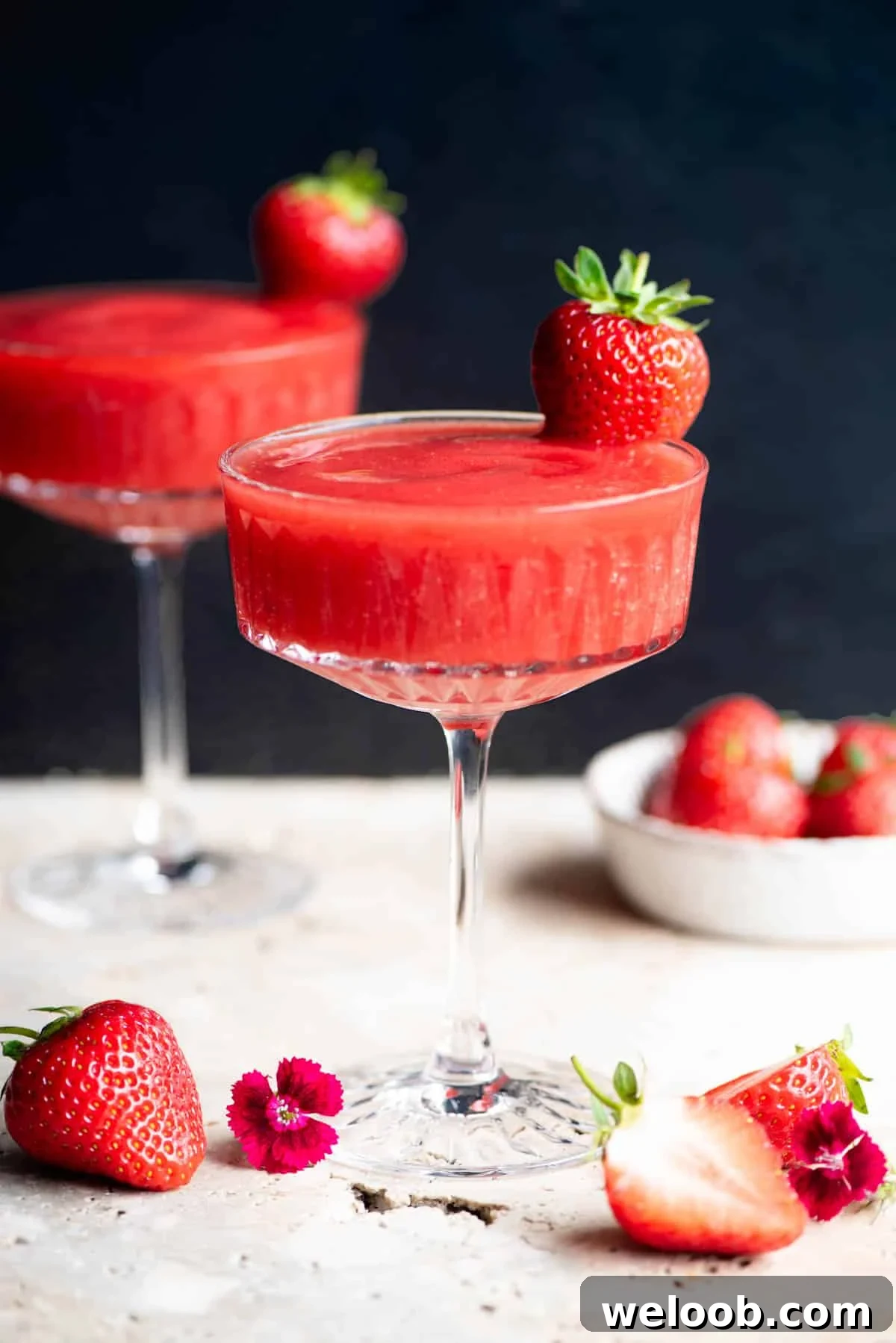 A side shot of a frozen strawberry cocktail in a glass garnished with a strawberry, the background is black.