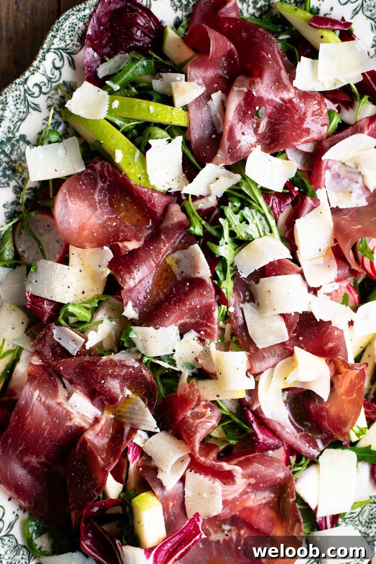 A close up of a salad on a green and white serving plate with bresaola, arugula, radicchio, radish and apple.