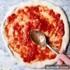 A close up of a pizza base getting tomato sauce spread on top before baking.