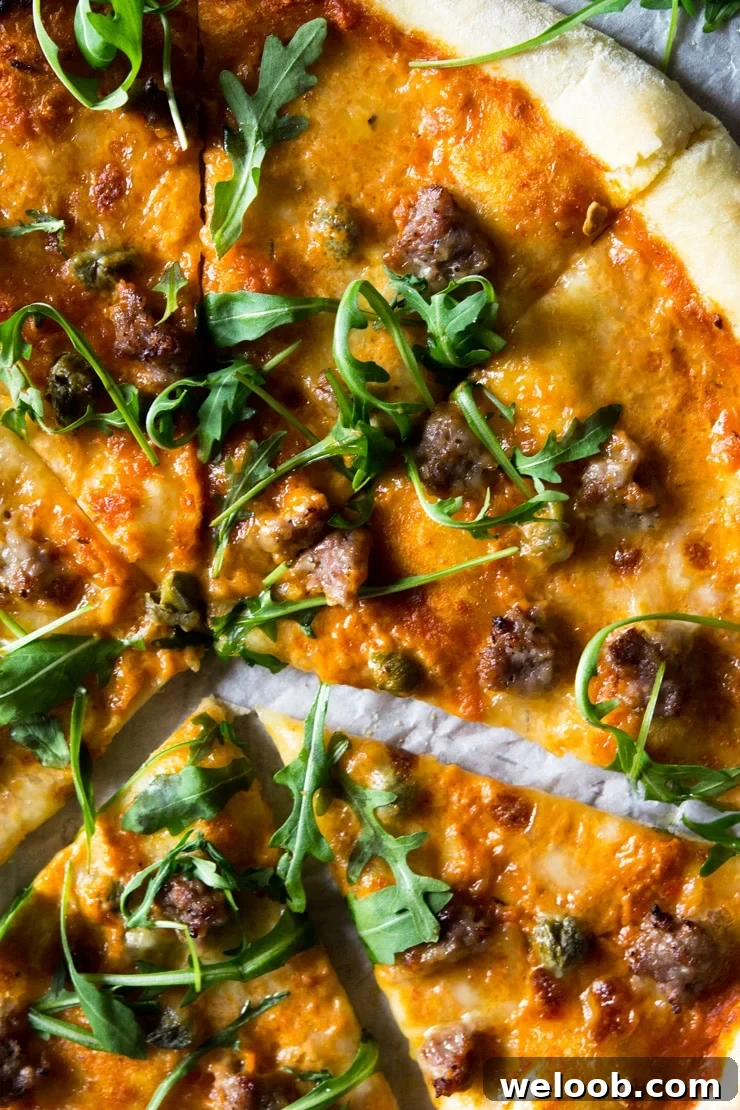 A close up of pizza with sausage and roasted red pepper sauce, melted mozzarella and topped with arugula.