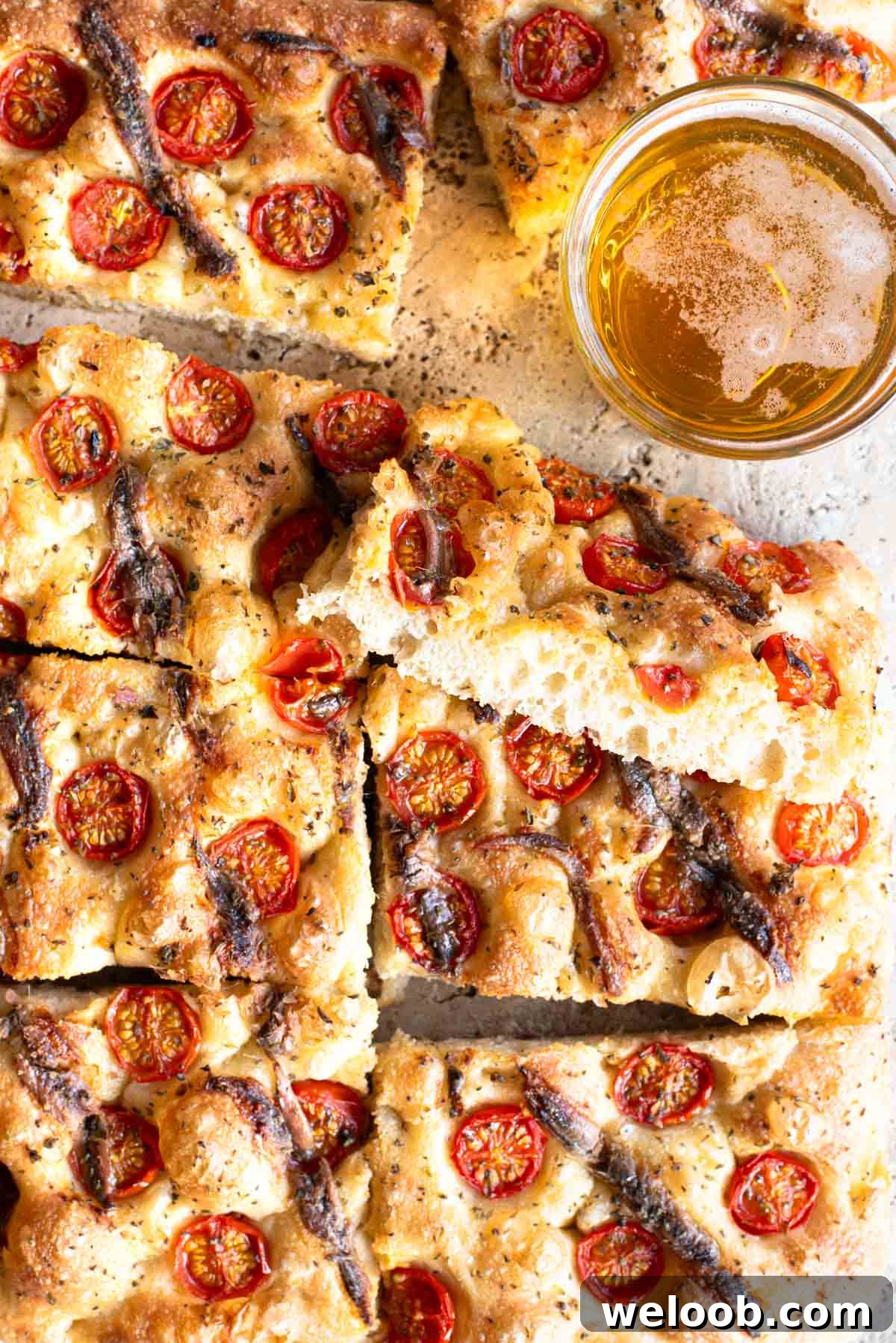 An overhead shot of a cherry tomato and anchovy focaccia cut into slices with a glass of beer at the side.