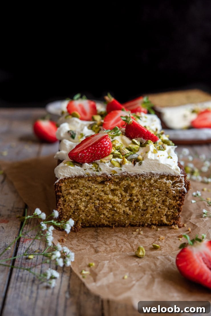 A side shot of a cut pistachio cake sitting on a wooden surface with strawberries and cream