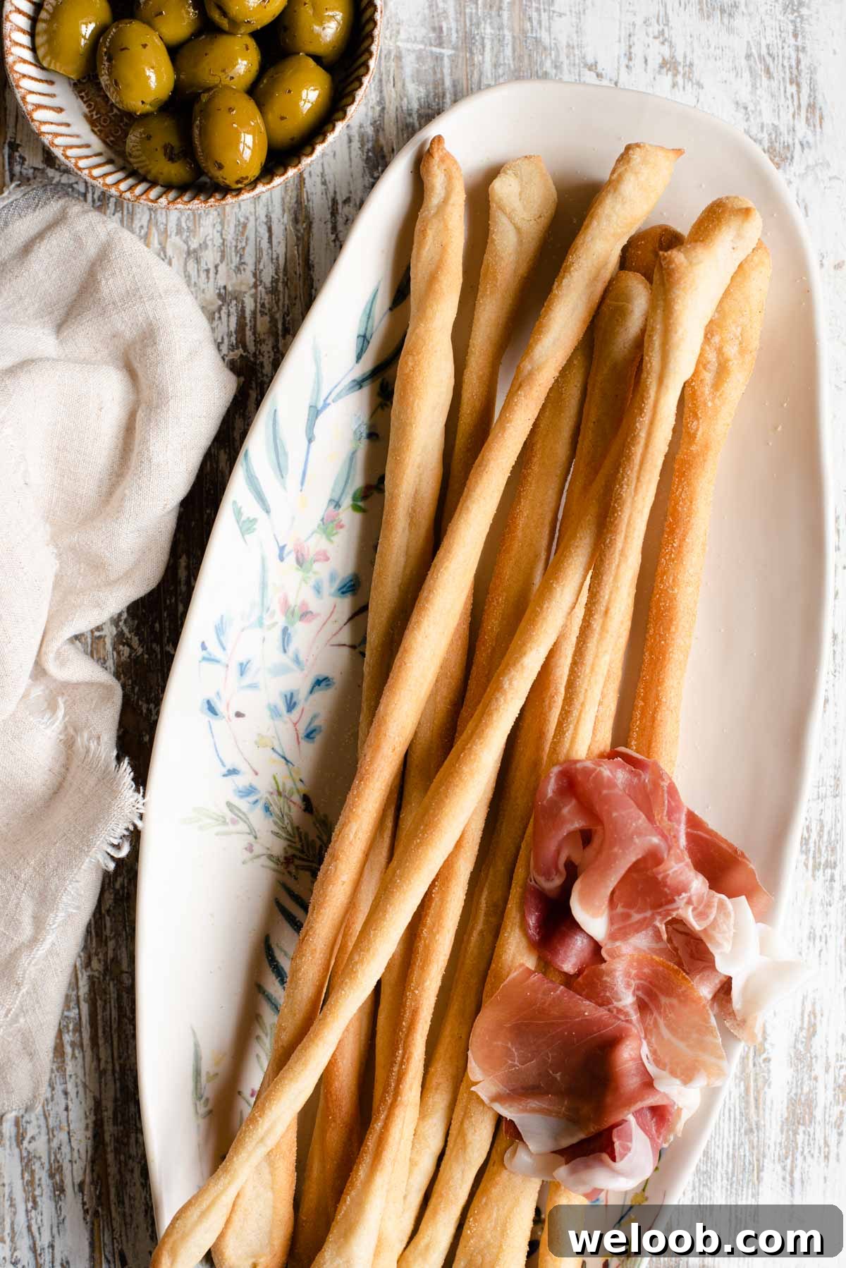 An overhead shot of Italian breadsticks on a serving plate with olives and prosciutto