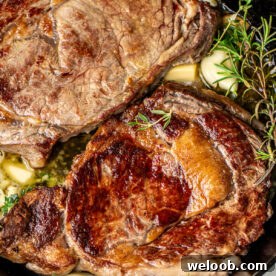 Two ribeye steaks in a cast iron skillet with rosemary, thyme, and garlic.