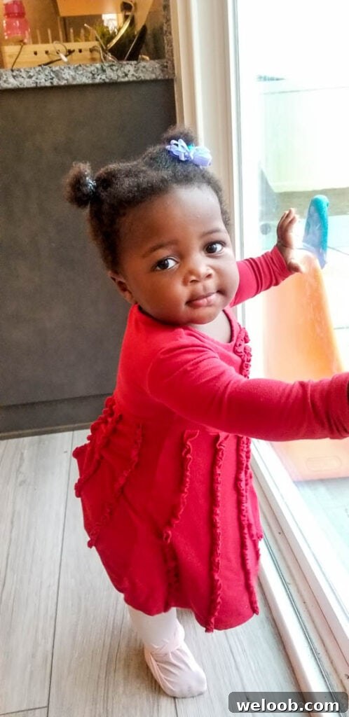 Baby in a pink dress standing at a window, looking at the camera.