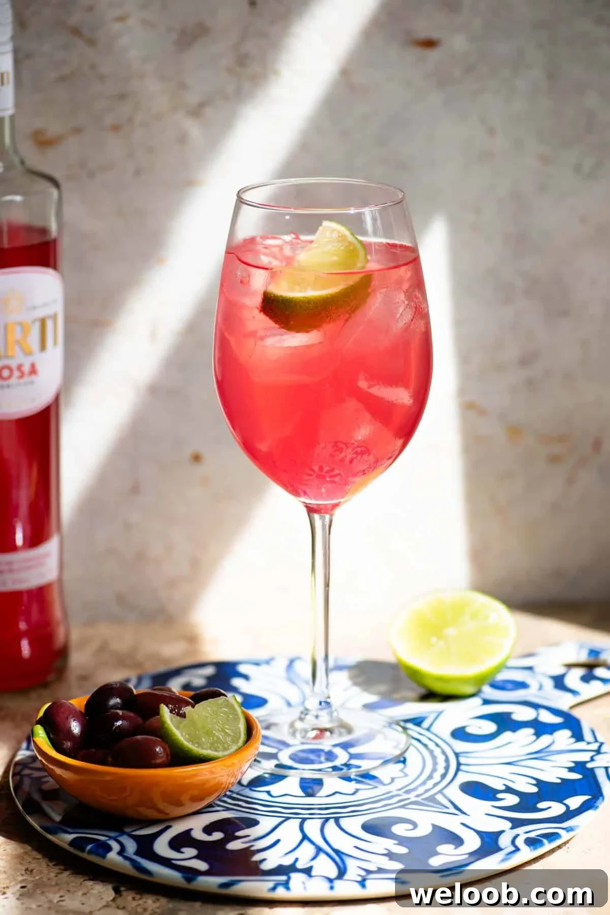 A Sarti Spritz in a wine glass with ice and a slice of lime sitting on a blue ceramic serving board with olive at the side.