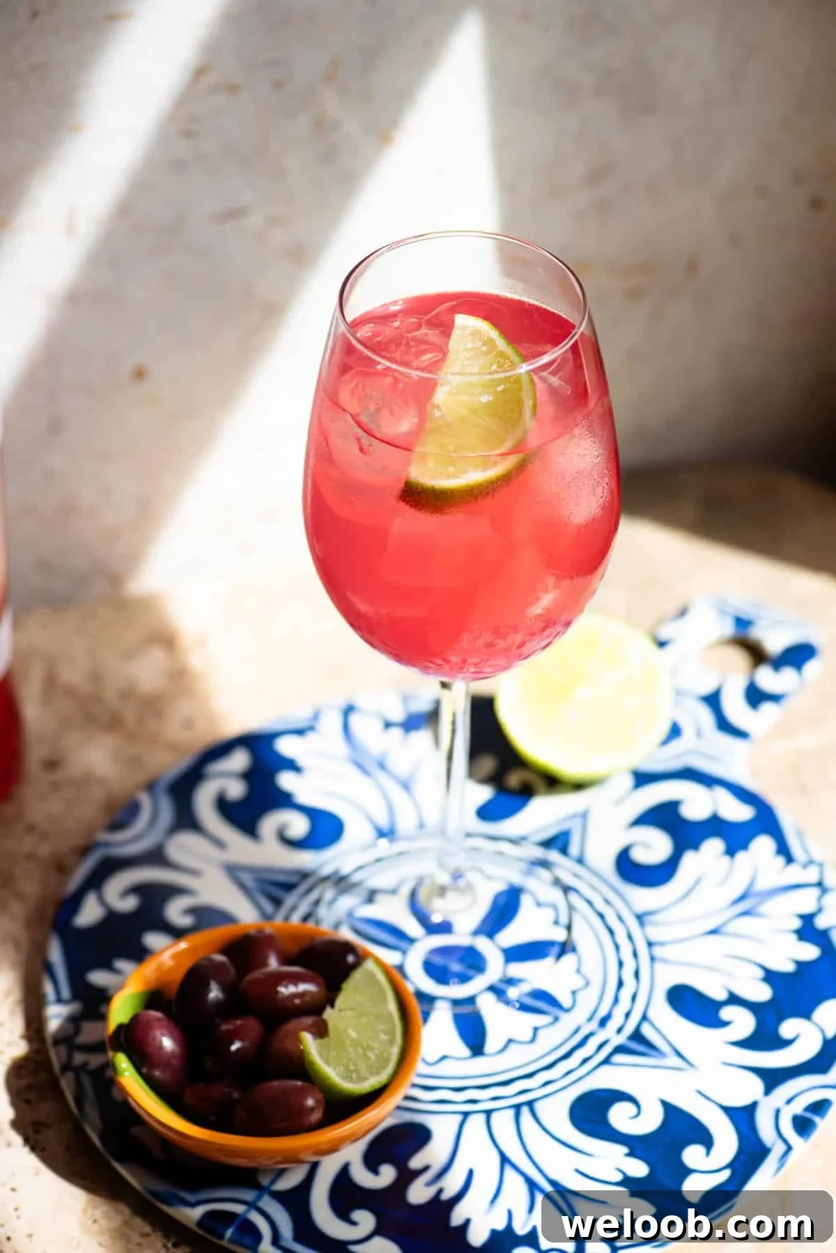 A close up of a sarti spritz with a wedge of lime on a blue patterned ceramic board with olives at the side.