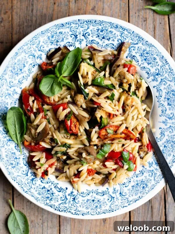 An orzo pasta salad with grilled veg in a blue bowl with a serving spoon at the side. The background is rustic wood.