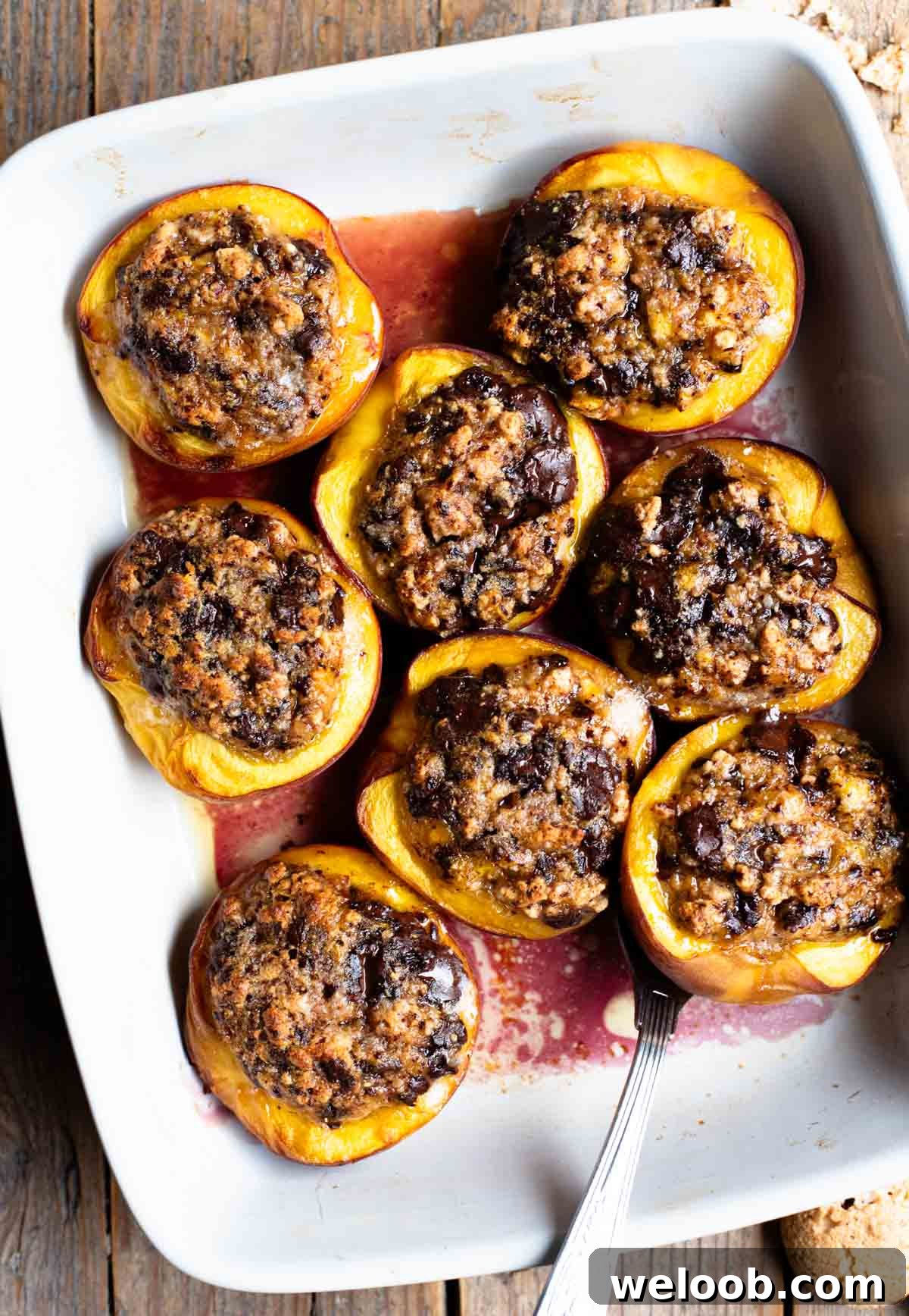 Piedmontese Baked Stuffed Peaches Recipe 2 An overhead shot of baked peaches stuffed with amaretti cookies and chocolate in a white baking dish on a rustic wooden background.