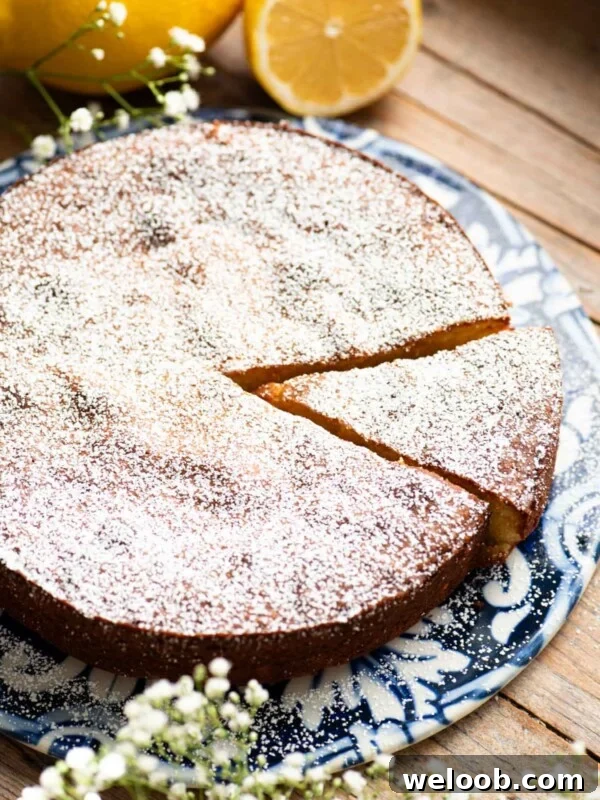 A white torta caprese on a blue ceramic board with fresh lemons behind it and small white flowers around.