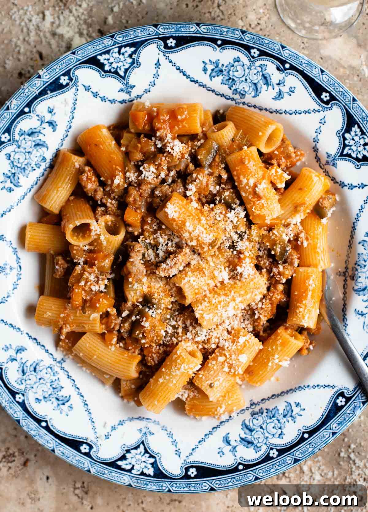 An overhead shot of rigatoni pasta with a sausage and eggplant ragu in a blue and white bowl on a travertine surface.