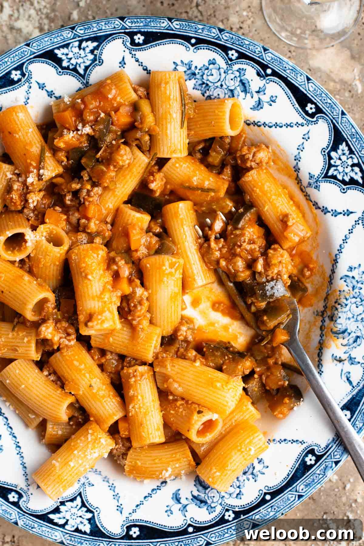 A close up of rigatoni pasta with a sausage and eggplant ragu in a blue and white bowl.