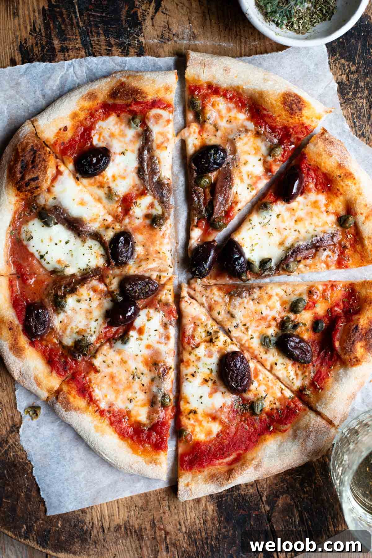 Mediterranean Olive, Anchovy and Caper Pizza with Oregano 2 An overhead shot of a pizza cut into slices on a rustic wooden board. The pizza is topped with olives, anchovies and capers.