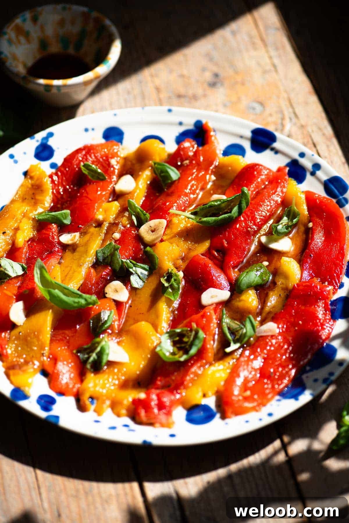 How to Make Italian Marinated Roasted Bell Peppers 4 Italian marinated roasted peppers on a blue and white plate topped with garlic and basil. The background is rustic wood.